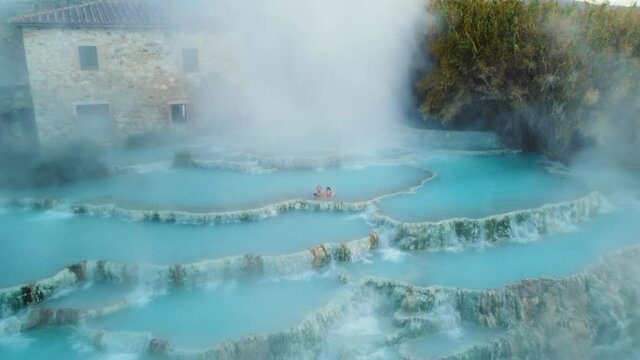 A young couple at the geothermal thermal hot springs bath and waterfall at Saturnia, Tuscany Italy close to Siena and Grosseto. Aerial drone at Cascate del Mulino, scenic and famous place in 4K UHD.