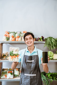 Asian Male Greengrocer Showing Vegetables And Showing A Phone Screen