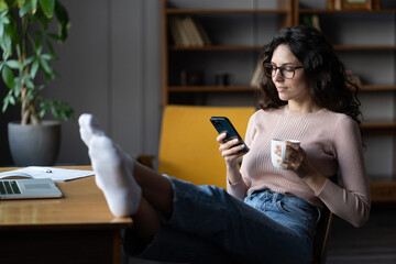Smiling millennial female employee procrastinate on workplace sit with cellphone and feet on desk....
