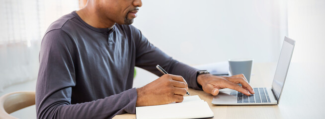Closeup black businessman hand holding pen writing on notebook, work on laptop computer in modern...