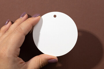 Fototapeta na wymiar Woman hand holding cardboard white blank tag of round shape with little hole for clothes in upper part in center of brown background. Tag mock up. Copy space.