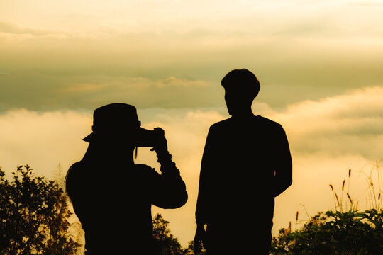 Silhouette of a cheerful couple traveler shooting pictures from a mobile phone. A woman photographs a man at sunrise over the misty and foggy morning. Man taking pictures of woman.