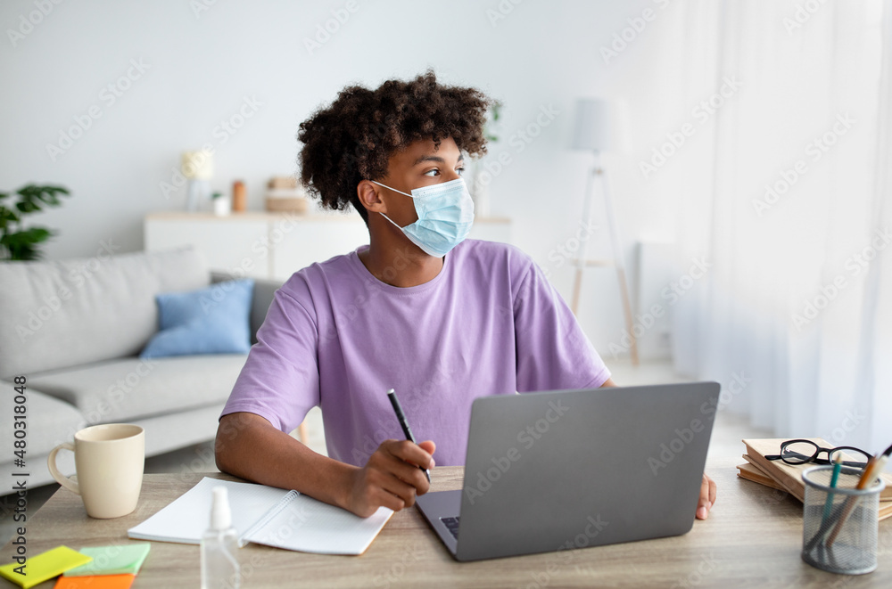 Wall mural black teenage student in face mask sitting at desk with laptop, taking notes in copybook, studying o