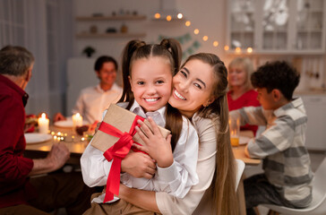 Happy young mother hugging her cute daughter with gift box during festive dinner at home