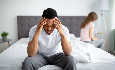 Fototapeta premium Erectile dysfunction, sex or relationship problems. Young black man sitting on bed, offended girlfriend on background