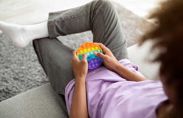 Unrecognizable black teenager playing with POP IT toy, using antistress sensory plaything, sitting on sofa at home - obrazy, fototapety, plakaty