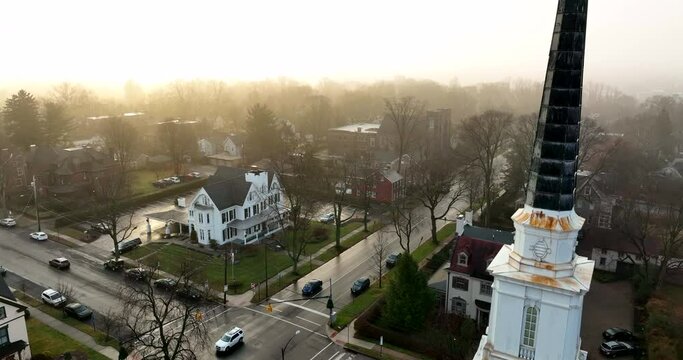 Rising aerial of church steeple in small town during dramatic morning light and fog. Winter scene.