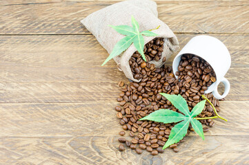 a composition of a coffee cup filled with coffee beans and a bag of coffee beans on a brown wooden background with green morijuana leaves. top view.
