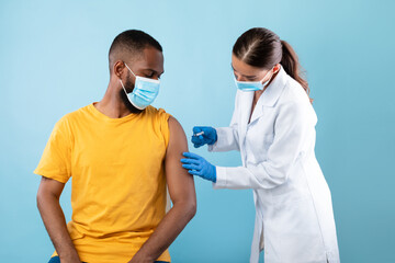 Millennial black man in protective mask receiving coronavirus vaccination on blue studio background