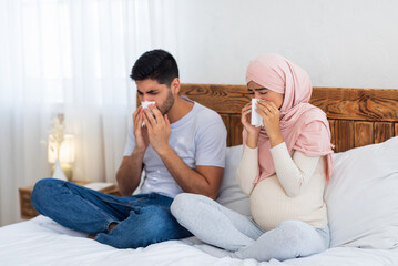 Young pregnant muslim couple got flu, having runny nose, sitting on bed at home, blowing nose...