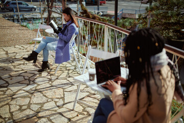 A young Asian woman sits in a cafe garden, reads a book, and relaxes while an African girl sits in the blurry foreground and typing on the laptop. Women having fun on their own.