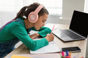 Girl in headset writing notes using laptop with empty screen