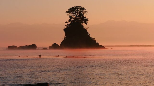 早朝の雨晴海岸より望む女岩とオレンジ色に染まる気嵐と背景の立山連峰