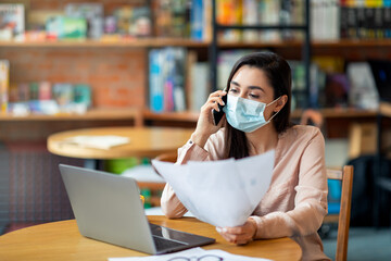 Busy arab woman freelancer in face mask talking on cellphone and checking papers, working on laptop computer in cafe