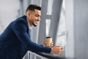 Happy Middle-Eastern Businessman Using Smartphone And Having Coffee While Waiting In Airport
