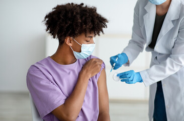 Medical doctor in rubber gloves giving covid-19 vaccine shot to Afro teenager at hospital....