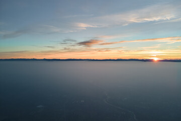 Aerial view of colorful sunset over white dense foggy clouds cover with distant dark silhouettes of mountain hills on horizon