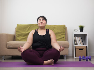 Young asian sport woman plus size sitting on the yoga mat practicing meditation. Fitness or exercise at home. Beautiful female relaxing after workout.