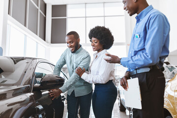 Car sales business. Manager talking to Afro couple, showing them new auto at dealership shop