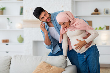 Pregnant muslim woman having prenatal contractions, caring husband calling doctor on smartphone, free space