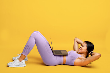 Online gym. Fit african american lady doing abs exercise sit-ups, training with laptop, exercising on yellow background