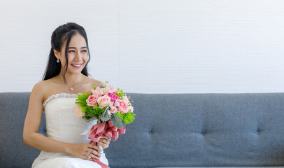 Portrait studio shot of Asian young happy cheerful beautiful female bride in white wedding dress with earrings and necklace sitting smiling look at camera on gray sofa hold pink roses flower bouquet