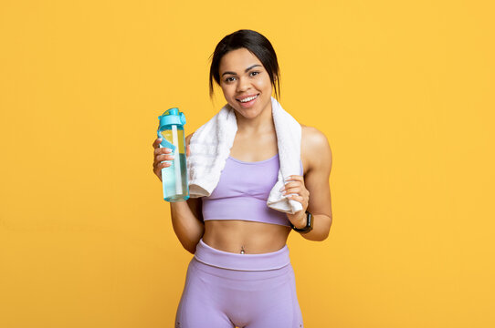 Stay hydrated during workout. Happy african american woman with towel and bottle of water on yellow background
