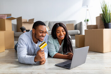 Loving black couple with laptop and credit card choosing furniture after moving to new home, free space