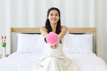 Obraz na płótnie Canvas Portrait shot of Asian young beautiful happy cheerful female bride in white long strapless wedding dress and veil sitting on bed smiling look at camera holding showing pink roses flower bouquet