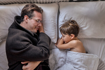 Father come to say goodbye to child boy before leave for work in morning, on bed. top view on sleepy man and kid at home, adult male in coat, in eyeglasses. family, monday, lifestyle concept