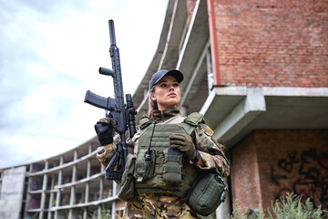 confident military woman with weapon, female in camouflage suit with rifle gun stand in grass field alone, abandoned building in the background. attractive female in gear, look at side posing - obrazy, fototapety, plakaty