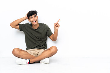 Young Argentinian man sitting on the floor surprised and pointing finger to the side