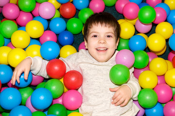 Fototapeta na wymiar A handsome boy of 4 years old lies among the plastic multi-colored balls in the playroom, smiling and looking at the camera, view from above