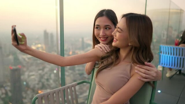 Asian woman friends in meeting and using smartphone selfie together at skyscraper rooftop restaurant in metropolis at summer sunset. Female friend enjoy outdoor lifestyle activity in the city at night
