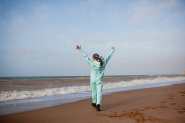 Teenager female walking outdoors in the beach in spring weather with wind, look to the sea, Girl dreased in blue hoody sweater and trousers