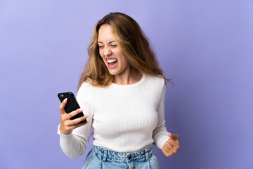 Fototapeta premium Young blonde woman isolated on purple background using mobile phone and doing victory gesture