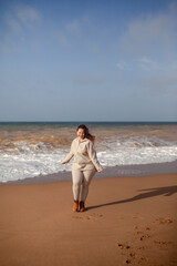 Beautiful woman wearing casual clothes, biege leggings, walking in windy sprin cold weather in the beach against the ocean.  Overweight woman have fun and enjoy the mood of view