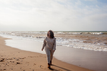 Beautiful woman wearing casual clothes, biege leggings, walking in windy sprin cold weather in the beach against the ocean.  Overweight woman have fun and enjoy the mood of view