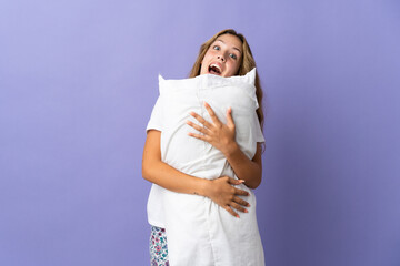 Young blonde woman isolated on purple background in pajamas and holding a pillow with happy expression