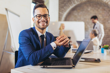 Happy businessman using laptop with team discussing project in the background.