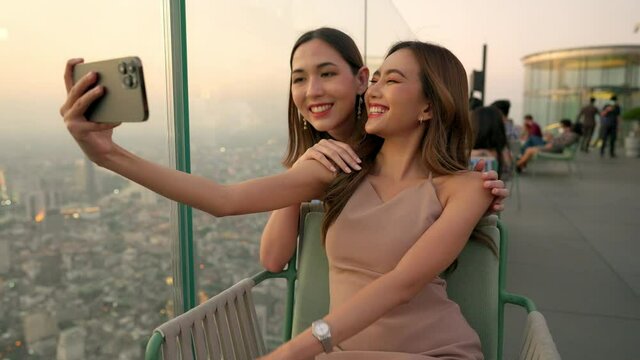 Asian woman friends in meeting and using smartphone selfie together at skyscraper rooftop restaurant in metropolis at summer sunset. Female friend enjoy outdoor lifestyle activity in the city at night
