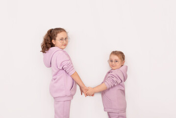 Two little beautiful smiling girls in fashionable spring clothes with a hood and glasses are holding hands. Carefree children in identical hoodies pose on a white background.