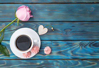 Black coffee in a white cup and meringue cookies on blue aged boards with orange streaks. Pink rose in the background. Top view, space for text