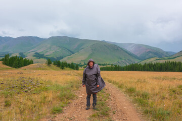 Satisfied tourist returns from mountains in overcast. Happy man in raincoat walks through hills and forest in bad weather. Traveler goes towards adventure. Hiker and mountain range under cloudy sky.