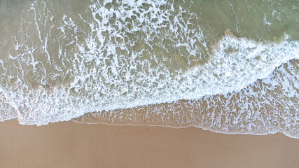 Aerial drone view perpendicular to the blue waves crashing on the sandy beach at the coast. The impact with wet wax creates white foam. The winter beach is empty. High quality photo