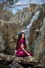 Naklejka na ściany i meble a brunette with long hair walks against the backdrop of a waterfall, a waterfall in the mountains, a photo session in a fabulous style