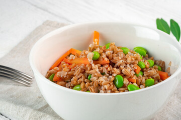 Vegan dish pearl barley with vegetables in a white bowl on a wooden table. Steamed pearl barley porridge with carrots, beans and pea sprouts, close-up - obrazy, fototapety, plakaty