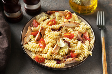 Pasta salad with grilled chicken, tomatoes and olive oil in a plate on a dark culinary background. A traditional Italian dish is girandole or fusilli with fried poultry fillet and vegetables