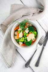 Mixed steamed vegetables in a white ceramic bowl. Vegan food - broccoli, carrots, beans, cauliflower and peas, top view