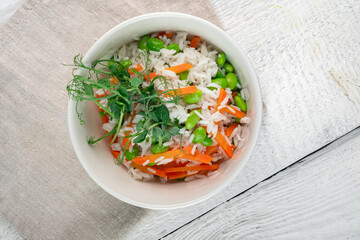 Vegan dish rice with vegetables in a white bowl on a wooden table. Steamed basmati rice with carrots, beans and pea sprouts, top view